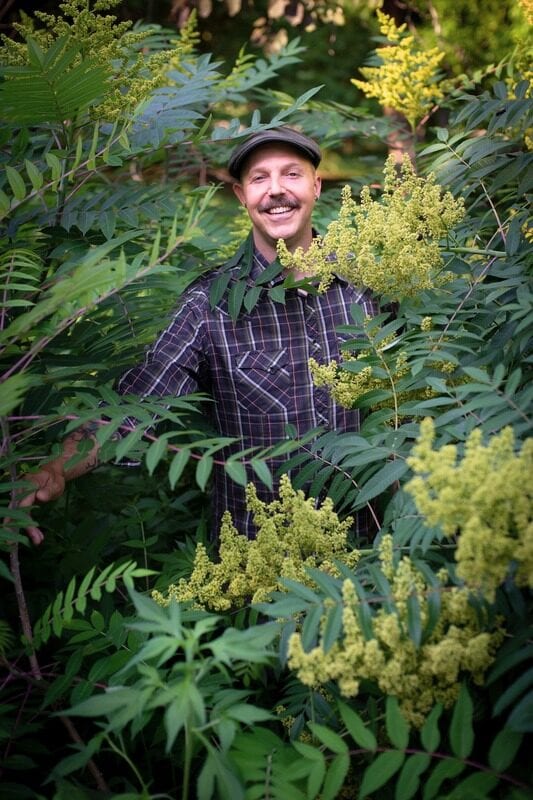Dan Pearson, smiling in a flat cap and plaid shirt, standing in a dense stand of flowering sumac